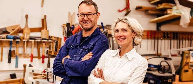 Ein Mann in Arbeitskleidung und eine Frau in Bluse stehen Rücken an Rücken in einer Holzwerkstatt und lachen.